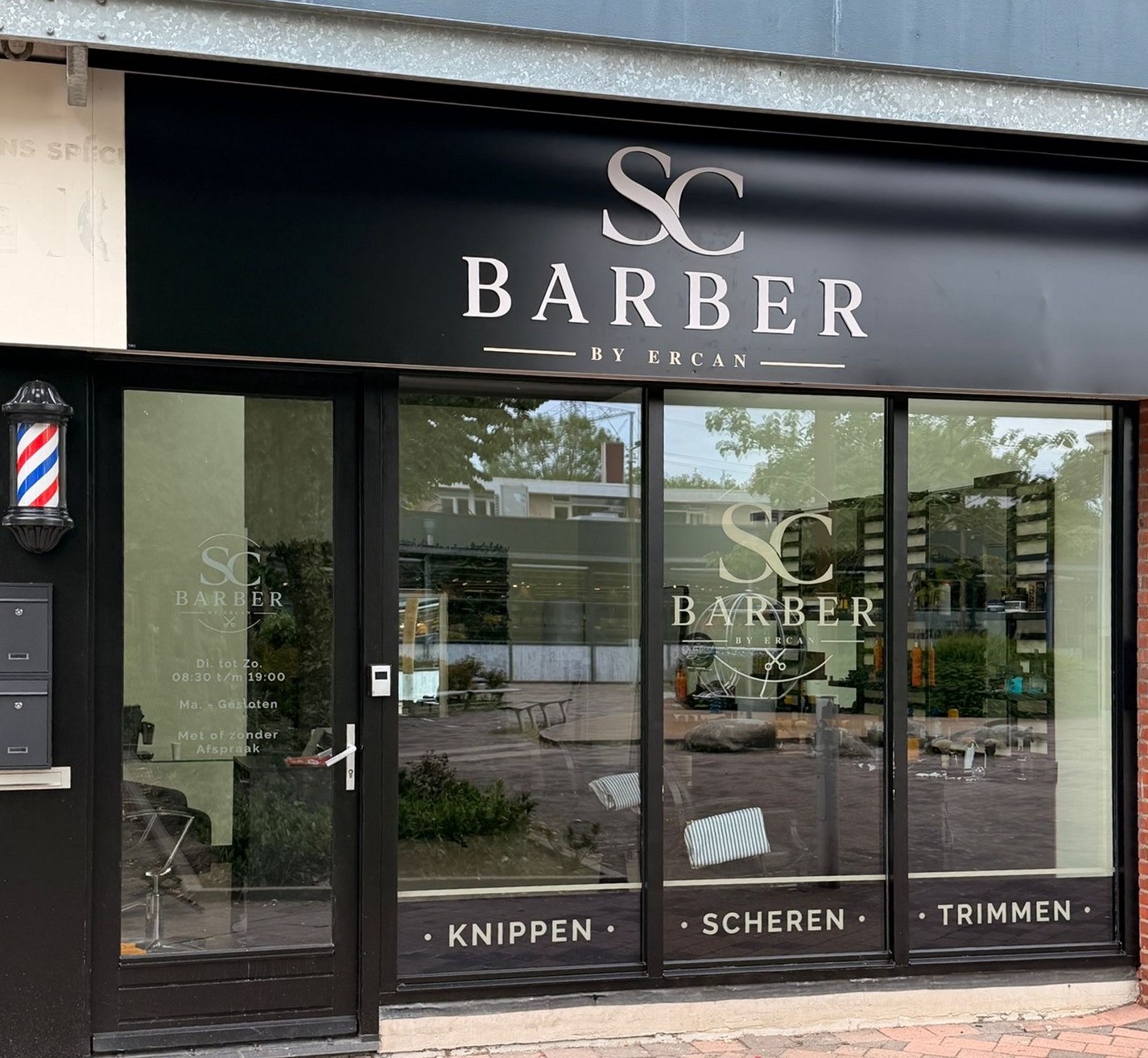De gevel van SC Barber: zwart uithangbord met SC Barber by Ercan, een kapperspaal en de raamletters knippen, scheren, trimmen