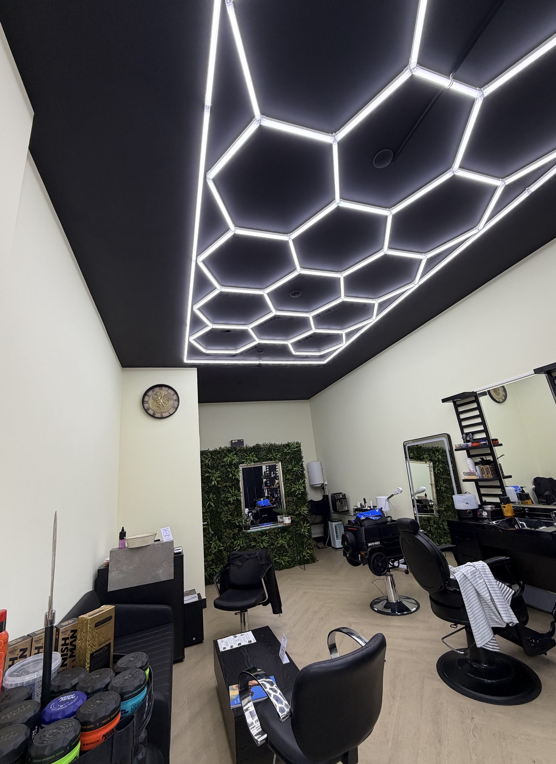 Interieur van SC Barber: verlicht plafond, zwarte kappersstoelen, spiegels en een plantenwand achterin